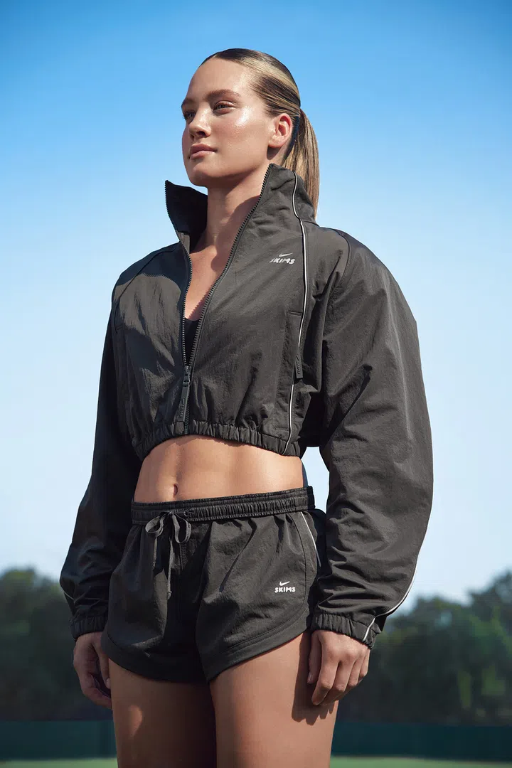 Nike与Kim Kardashian创立的SKIMS携手推出NikeSKIMS 2026秋季系列，画面中一名金发女模特儿站在户外，头发梳成利落的低马尾，身穿深炭灰色短版拉链运动外套及同色系运动短裤。外套采用高领设计、宽松长袖与弹性收腰剪裁，正面饰有NikeSKIMS白色品牌标志，短裤则搭配抽绳腰头与白色滚边细节。模特儿露出腰腹线条，背景为晴朗的蓝天及远处的绿色树木，展现NikeSKIMS将运动机能、女性身形剪裁与日常时尚穿搭结合的设计理念。