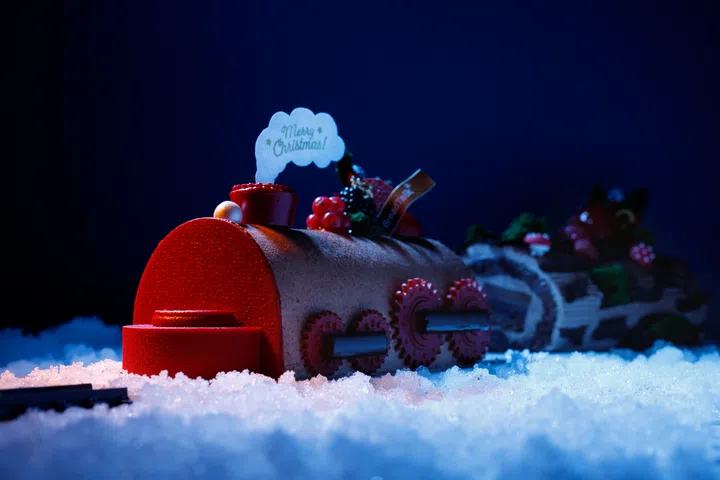 BreadTalk 的红色圣诞火车造型 logcake，配有齿轮细节、浆果点缀与“Merry Christmas”装饰，置于蓝色灯光下的雪景中，呈现浓烈节庆氛围。2025 新加坡圣诞蛋糕推荐｜好吃又好看的 Christmas Cakes & Logcakes 精选清单