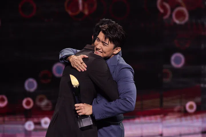 2025年红星大奖颁奖典礼上，张哲通身穿深蓝色西装，在舞台上拥抱另一位艺人，脸上露出开心笑容，手中握着奖座，背景是红色灯光点缀的颁奖舞台。