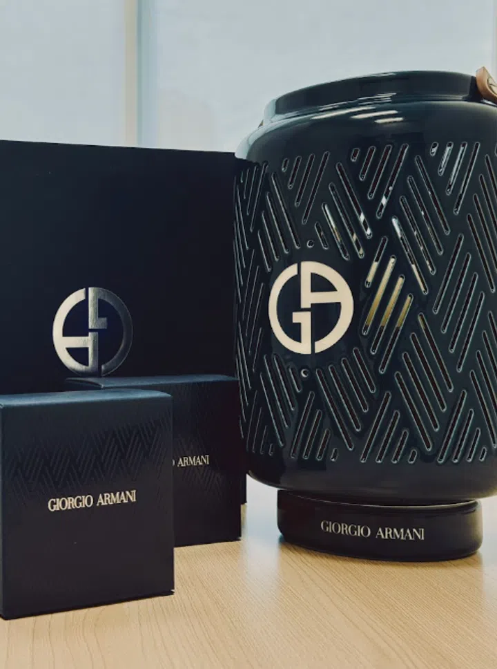 Giorgio Armani中秋礼赠陈列于浅木色桌面，右侧为深色圆筒状灯笼造型礼品，表面布满交错的斜线镂空纹样，中央饰有醒目的金色GA标志，圆形底座印有品牌全名。左侧配套深色方盒带有细密几何纹样及金色字样，后方竖立的深色包装上也饰有大型GA标志。