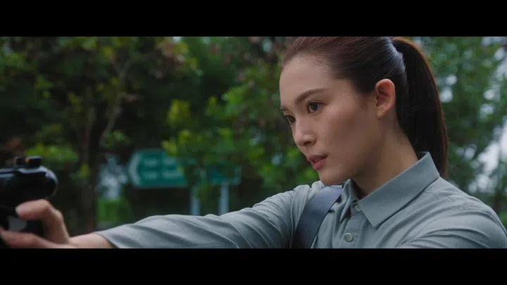 周智慧在电视剧中的一幕动作场景，扎着高马尾的她神情专注，侧脸入镜，举枪瞄准前方。她身穿灰色衬衫，造型干练利落，眼神坚定而警觉，呈现出紧张的对峙氛围。背景是略微虚化的树林与路牌，暗示户外追踪或行动场景，整体画面色调偏冷，强化剧情的悬疑与紧迫感。红星大奖2026《最佳女主角》入围者