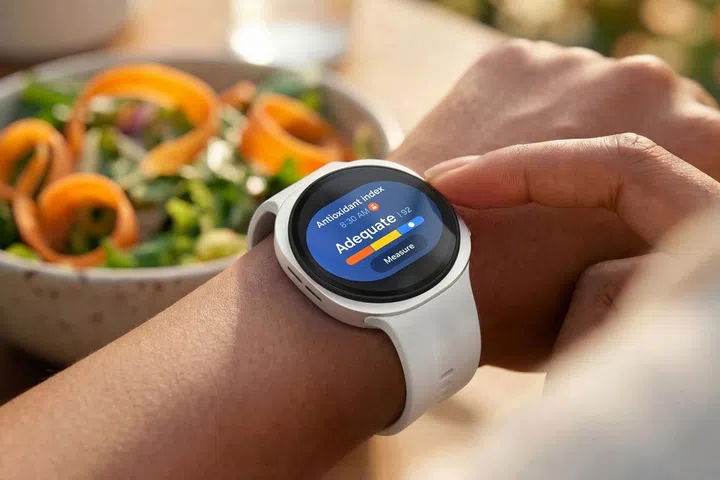 一名用户佩戴白色表带的Samsung Galaxy Watch9，正在查看手表上的Antioxidant Index（抗氧化指数）功能。圆形表盘显示抗氧化指数为“Adequate（充足）”，并以彩色指标条呈现测量结果，下方设有“Measure（测量）”按钮。用户另一只手轻触手表边缘，准备进行操作。背景摆放一碗色彩丰富的沙拉，包含绿叶蔬菜及橙色蔬菜丝，在温暖的自然光下营造健康饮食与日常生活场景，突显Galaxy Watch9可协助用户追踪健康数据，并结合饮食与生活习惯进行日常健康管理。