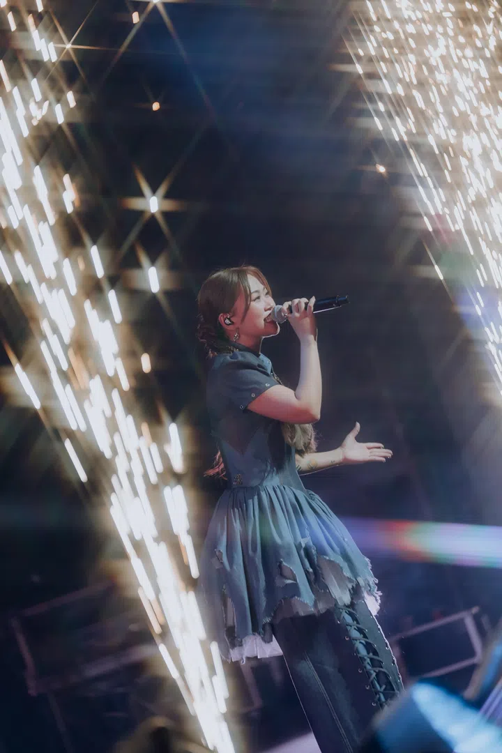 DIOR大颖在新加坡演唱会现场侧身演唱，身穿蓝色层次感舞台短裙搭配长裤造型，手持麦克风投入演唱，一手自然延展与观众互动。舞台两侧灯光如流星般倾泻而下，形成闪耀光束效果，整体画面充满动感与情绪张力，展现她真挚而具感染力的现场演唱魅力。