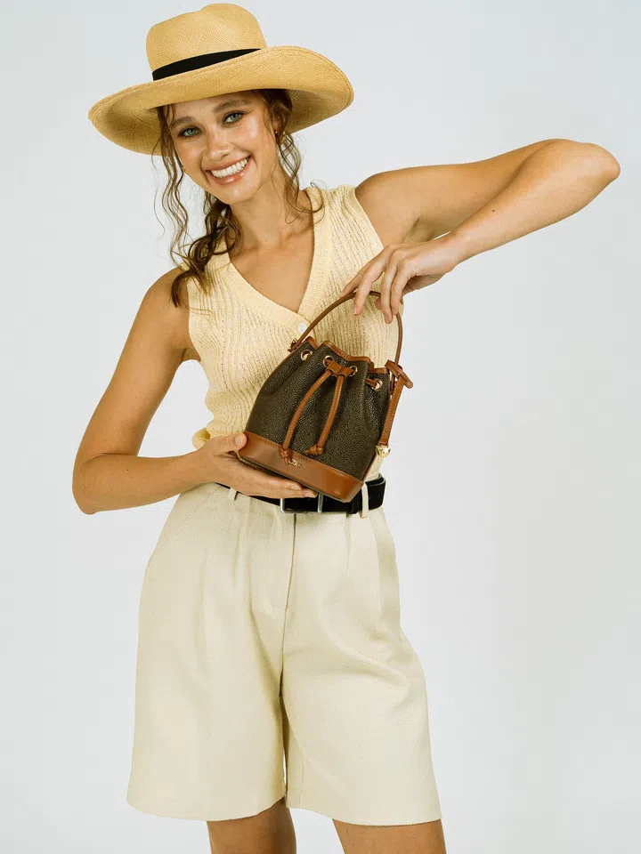 模特手提 Tocco Toscano Heritage Bucket Bag Mini 水桶包，以米色针织上衣、宽松短裤与草帽打造轻松复古穿搭风格。包款采用深棕与焦糖色拼接设计，搭配经典抽绳与金属细节，呈现品牌标志性的复古优雅感。整体造型带有浓浓度假氛围，同时展现水桶包轻巧实用、适合日常搭配的特点，非常适合作为母亲节送给妈妈的时尚包包礼物。母亲节礼物推荐2026：从实用家居到时尚科技，这些礼物妈妈真的超爱！