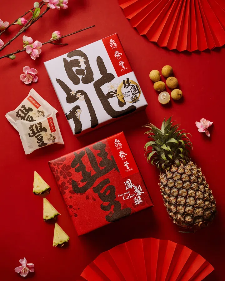 画面展示鼎泰丰（Din Tai Fung）农历新年限定凤梨酥与桂圆酥礼盒平铺在红色背景上，整体构图以对称方式呈现浓厚新春氛围。上方为白色包装的桂圆酥礼盒，盒面以书法字体与金色花纹点缀，旁边摆放完整桂圆与剥开的桂圆果肉，呼应口味来源；左侧可见独立包装的桂圆酥小包装。下方为红色包装的凤梨酥礼盒，盒面同样以书法字样设计，旁边摆放整颗凤梨与切块凤梨果肉，突出凤梨酥的主要食材。画面四周点缀粉色梅花枝与红色纸扇装饰，加强节庆视觉元素，整体呈现传统中式美感与节日送礼氛围，适合新春拜年与送礼选择。文章标题：“2026马年年饼推荐：新春送礼与拜年点心精选”。