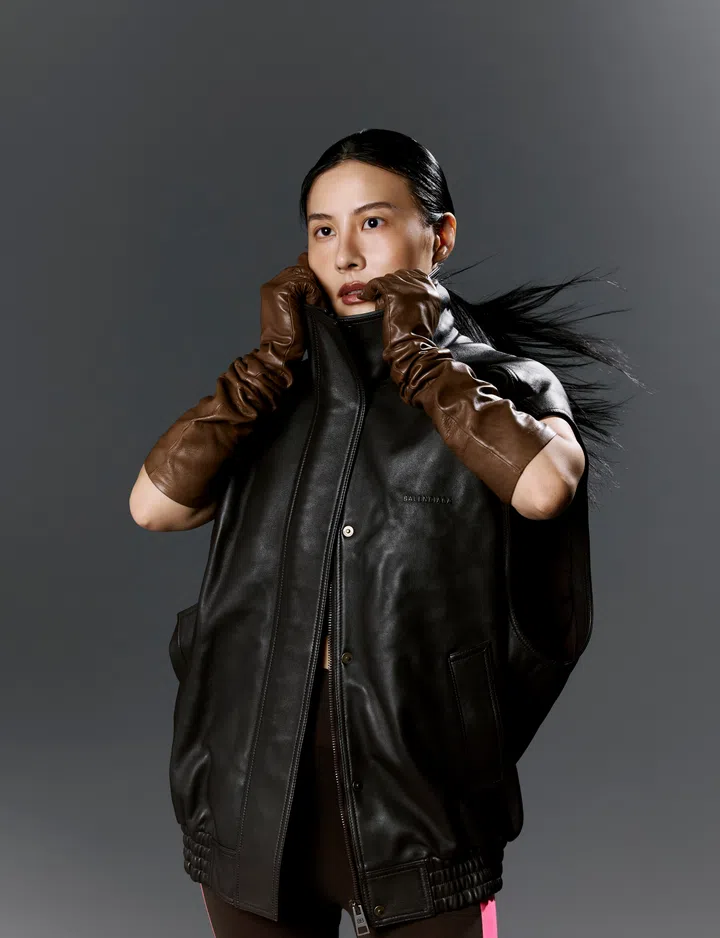 Savina Chow站在深灰色摄影棚背景前，身穿Balenciaga黑色皮革无袖外套，搭配咖啡棕色长皮手套，双手轻轻拉起高领外套遮住下半张脸，露出坚定而专注的眼神。她将黑色长发梳成低马尾，发尾随风向右飘扬，为整体造型增添动态感。皮革外套采用宽松Oversized剪裁，两侧设有大型口袋，整体造型简约俐落又充满未来感，展现高端时尚品牌标志性的先锋美学。摄影采用柔和棚灯照明，突显皮革材质的细腻光泽及人物立体五官，整体氛围冷静、自信且富有力量。