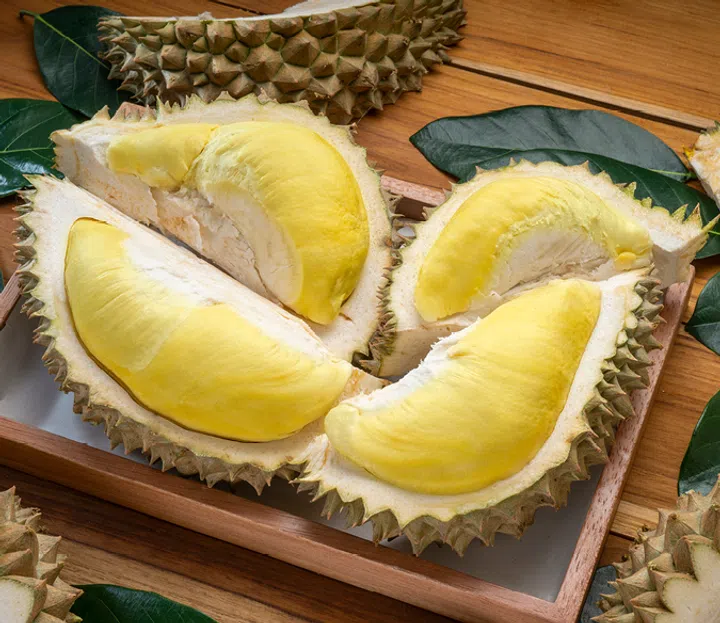 打开的榴莲放在木盘上，果肉金黄饱满，周围散落着榴莲壳和绿色叶子，背景是木质桌面。