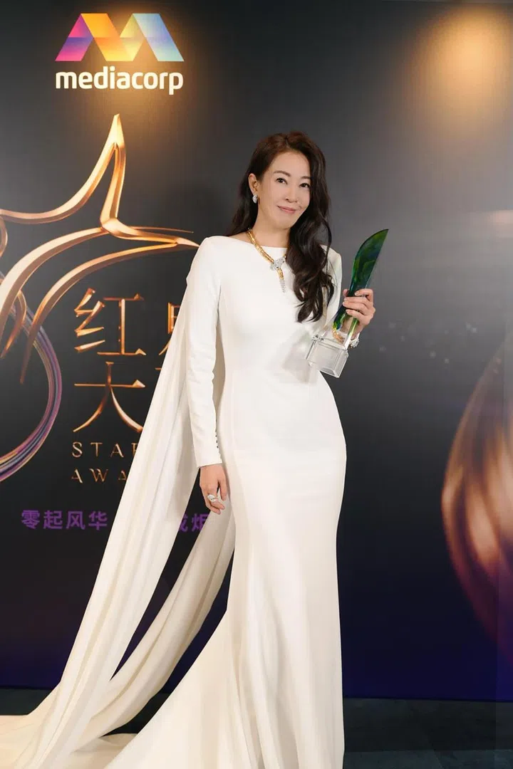 这是一张在《红星大奖》活动现场拍摄的人物照片。刘子绚身穿一袭剪裁贴身的白色长礼服，整体线条简洁流畅，裙摆自然垂落，并在身后延伸出飘逸的披肩式拖尾，增添优雅气场。她手持绿色透明质感的奖座，面带从容自信的微笑，直视镜头，状态沉稳而温柔。
她的造型以低调精致为主，搭配简约的金色项链与耳环，妆容干净自然，长发微卷披肩，整体呈现出成熟大方的气质。背景为深色渐变的活动拍照墙，上方清晰可见“mediacorp”标志，旁边配有《红星大奖》的金色星形视觉元素与活动字样，营造出隆重正式的颁奖氛围。
画面右侧前景略微虚化，有观众或工作人员的身影，增加现场感与层次感。整张照片捕捉了她在得奖后的定格瞬间，既优雅又充满荣耀感。刘子绚首夺视后：22年7次提名终圆梦，她把时间走成答案