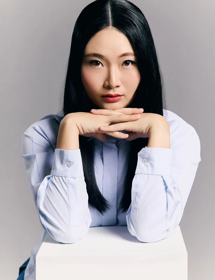 一张棚内拍摄的人物肖像照片：画面中央是一名长直黑发的年轻女性，身穿浅蓝色长袖衬衫，双手交叠托着下巴，身体微微前倾，靠在白色方形台座上，正面直视镜头。她的妆容自然，肤色明亮，表情沉稳而专注，带出理性与自信的气质。背景为简洁的浅灰色纯色布景，光线柔和均匀，整体画面干净、专业，具有杂志人物专题与编辑摄影风格，突显人物的专注神情与内敛力量。cloop founder tan yin ling cloop环保纺织品箱子二手衣