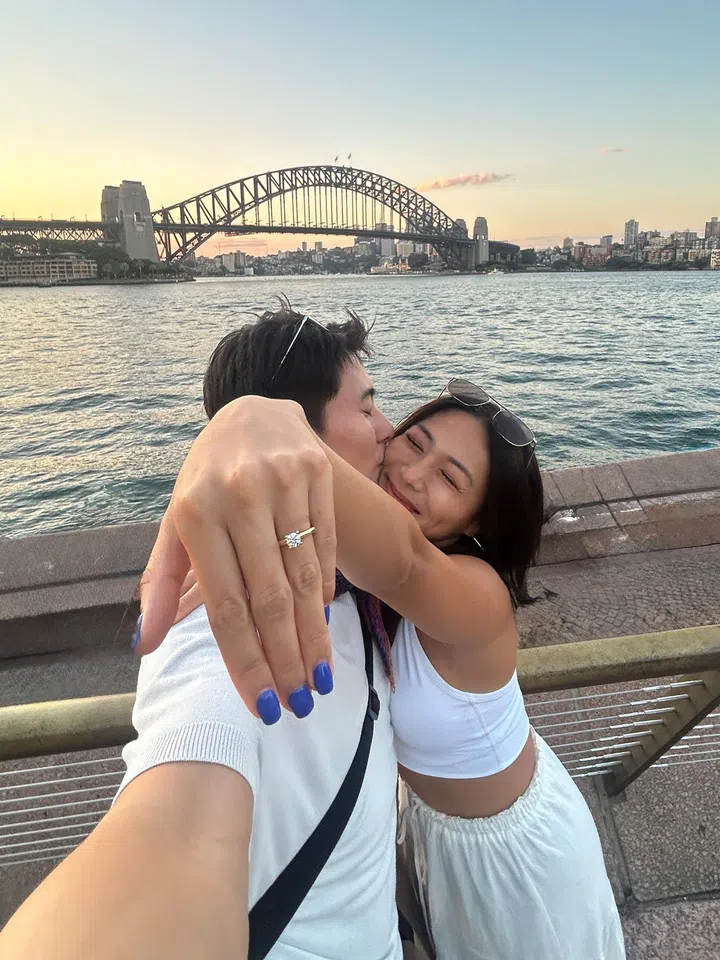 在澳洲悉尼海港大桥前的傍晚时分，一对情侣在海边栏杆旁亲密拥抱合影，女生伸出戴着订婚戒指的手对着镜头，脸上带着开心笑容，背景是夕阳下的港湾与城市天际线，画面洋溢幸福甜蜜氛围。Rachel Wan：最好的感情，是让我更像自己