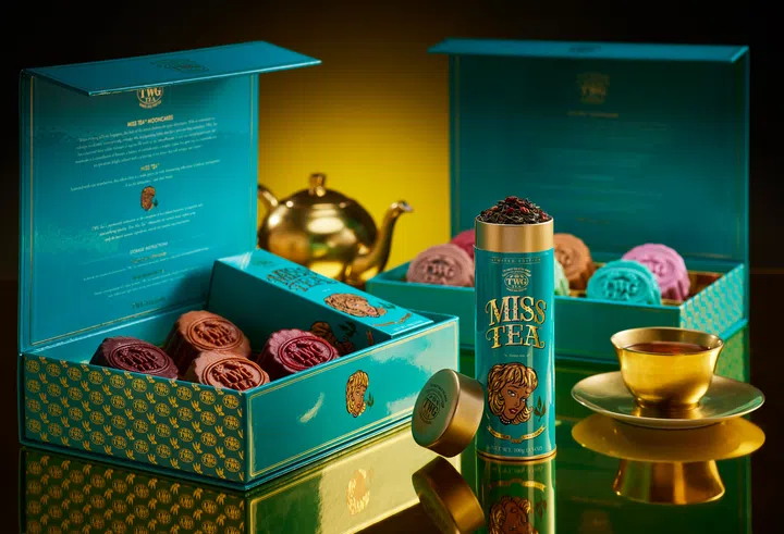 TWG Tea 2026中秋Miss Tea月饼系列场景图，以品牌标志性的蓝绿色礼盒为主视觉，前后两盒分别陈列深红、棕色、粉色、绿色及蓝绿色等不同颜色的月饼。中央摆放一罐Miss Tea限量茶叶，旁边配有金色茶壶与盛有茶汤的金色茶杯，整体以蓝绿与金色为主调，突出茶香月饼与品茶搭配的节庆氛围。