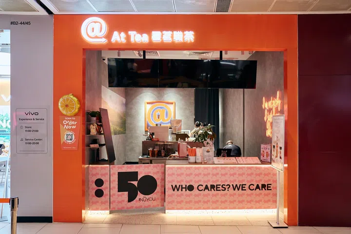At Tea位于313@somerset的门店外观，以橙色店面搭配《女友》50周年粉色联名设计，柜台印有“WHO CARES? WE CARE.”及《女友》50周年标志，展示《女友》与At Tea联名合作主题及限定饮品宣传。