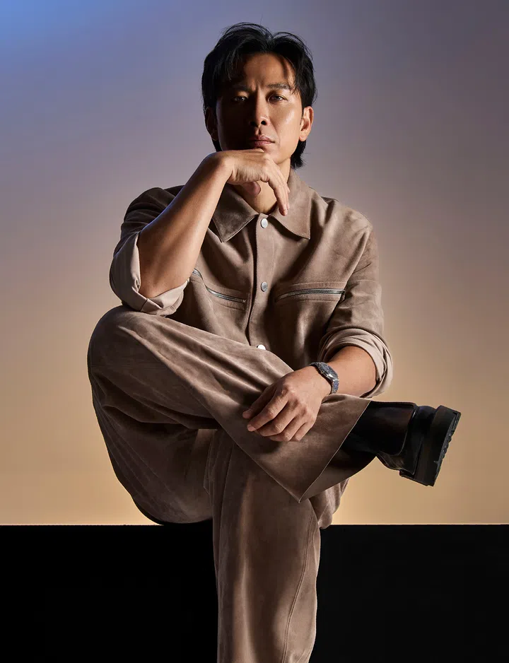 戚玉武身穿浅褐色麂皮套装，坐在昏黄光影中，以沉静姿态凝视镜头，展现成熟与思考的气质。