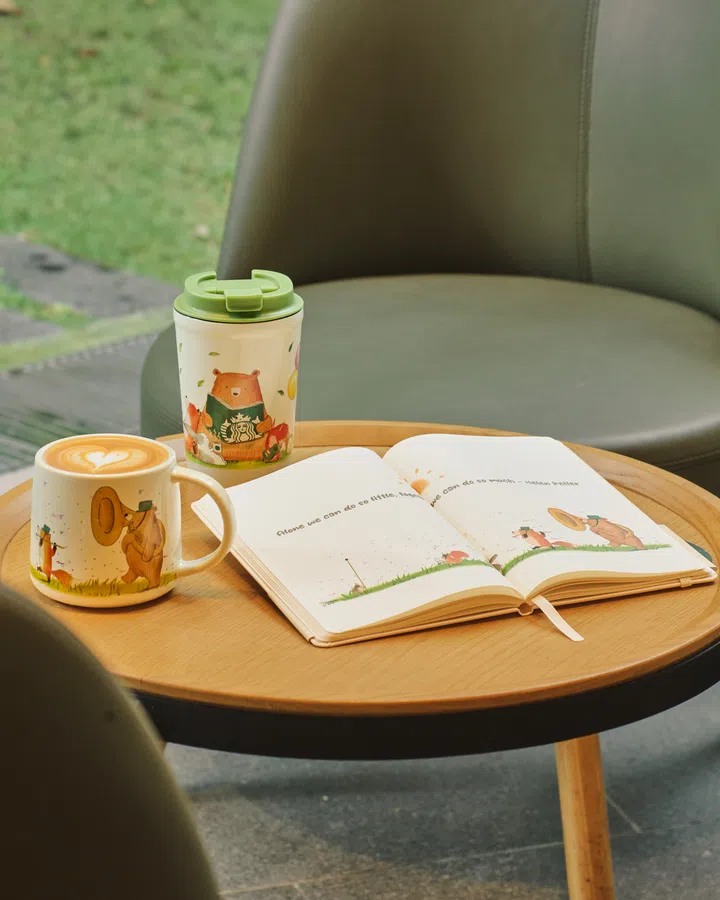 一张拍摄于咖啡馆的照片，木质圆桌上放着一杯拿铁、一只印有插画的马克杯、一只绿色杯盖的随行杯，以及一本打开的笔记本，整体氛围温暖宁静，充满惬意的午后感。