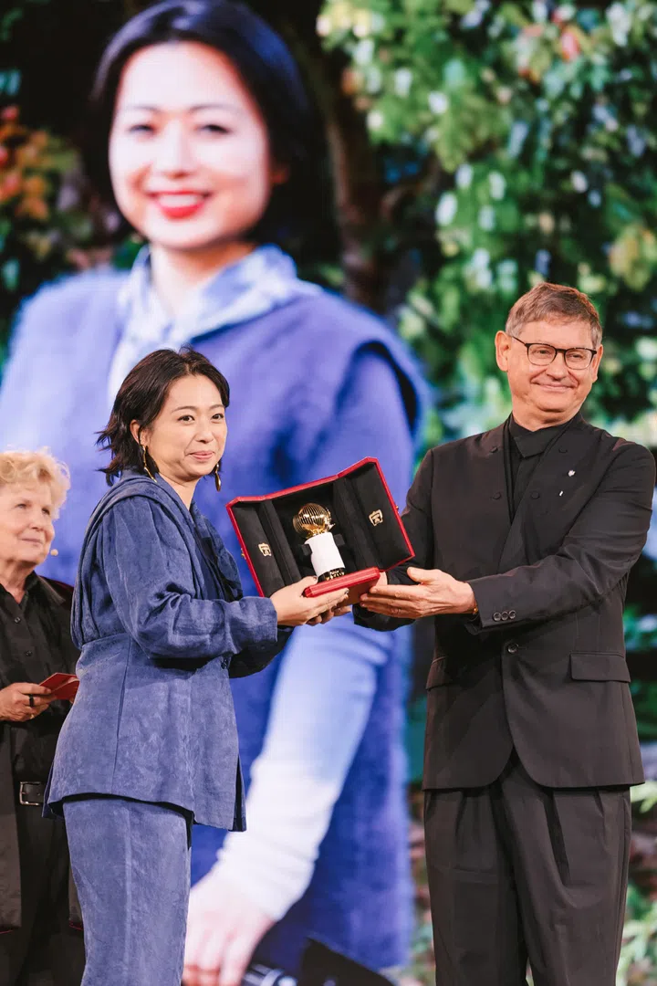 BloomGEM联合创办人兼首席执行官叶婷于Cartier Women’s Initiative 2026 Awards Ceremony上领奖，从Cartier文化与慈善事业主席Cyrille Vigneron手中接过象征荣誉的奖座。叶婷身穿蓝色套装，双手捧着打开的红色奖盒，盒内陈列着Cartier Women’s Initiative标志性的金色奖座。身后大型LED屏幕播放她的肖像照片，记录她获得2026年Cartier Women’s Initiative伙伴计划（Fellow）的重要时刻，肯定她以生物染色技术推动时尚与美妆产业迈向更环保、更永续发展的创新贡献。