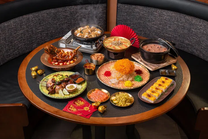 一桌丰盛的新春团圆宴摆放在圆形餐桌上，中央为寓意吉祥的鱼生拼盘，搭配各式中式佳肴，包括红烧整只肉类主菜、滋补汤品、砂锅料理、烤肉拼盘及精致点心等。桌面点缀红包与金元宝装饰，背景以红色折扇衬托节庆氛围，整体呈现适合农历新年期间与家人好友共享的丰富中式宴席与团圆聚餐场景。新加坡大年初一初二照常营业餐厅推荐：8家不打烊好去处（2026马年）