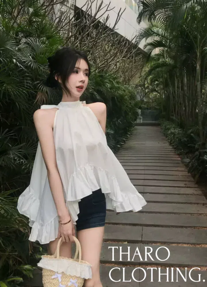THARO CLOTHING女装穿搭照。模特站在两侧绿植环绕的石板步道上，身穿白色挂脖无袖宽松上衣，领口带有细密褶皱，露出肩膀与手臂；衣身呈向外展开的A字轮廓，不规则下摆拼接宽幅荷叶边，形成前短、两侧较长的层次。下搭深蓝色牛仔短裤，一手提着原色编织手袋，袋口饰有白色花边，袋身点缀浅色小蝴蝶结。模特将黑发松松盘起，脸侧留有碎发，佩戴细手链，侧头望向画面右方。背景可见棕榈、灌木及向远处延伸的步道，右下角印有白色“THARO CLOTHING.”字样。