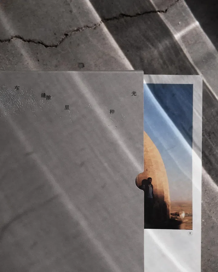 一本摄影文集的封面与内页叠放在地面上拍摄，灰色半透明封套上可见压印的书名文字“在 缝隙 里 种 光”，表面有细小凸点纹理，右侧露出内页照片的一角。背景为带有裂痕的水泥地面，斜射的阳光在地面与书页上形成明暗交错的光带。内页照片中可见一座拱形建筑与远方沙漠景色，一名人物站在拱门阴影中，整体画面呈现出光从缝隙与空间边缘流入的感觉，呼应书名主题。林道锦《谁在缝隙里种光》