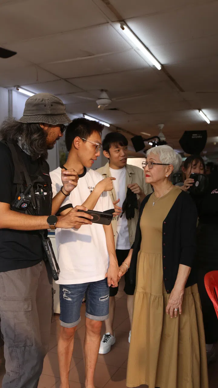 《百万红包 A Good Fortune》拍摄现场幕后花絮照，导演 Jason Lee 站在中间手持手机与演员讨论画面内容，身旁有工作人员与演员围在一起观看画面，一位白发年长女演员站在前方认真聆听指示，现场可见摄影与灯光设备，整体氛围专注而温和，呈现电影拍摄过程中导演与演员沟通、调整表演细节的真实片场瞬间，展现贺岁电影《百万红包》的幕后制作过程。