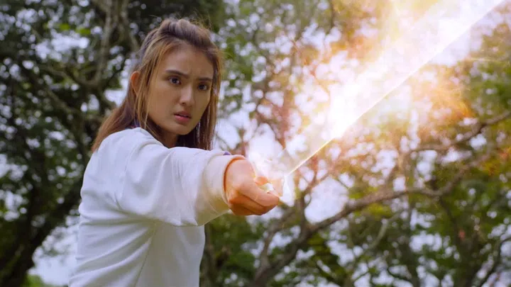 洪凌在《带剑女孩》中的一幕动作场景，身穿白色上衣的她神情紧绷，眼神坚定而警觉，手持发光的长剑直指前方。剑身散发出耀眼光芒，带有奇幻元素，增强视觉冲击力。画面背景为树林环境，阳光从树叶间透出，营造出紧张又带有神秘感的氛围。镜头捕捉她准备出击的瞬间，展现角色的力量感与决心。红星大奖2026《最佳女主角》入围者