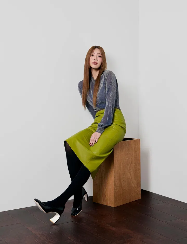 一位女性坐在木质方块上，背景为简洁的浅灰色墙面与深色木地板，整体画面干净利落。她留着顺直的长发，中分造型，妆容精致，神情从容自信，微微侧身面对镜头。她身穿带有细腻光泽感的灰色长袖上衣，搭配亮绿色高腰半裙、黑色丝袜与黑色高跟鞋，整体造型时尚利落又带有鲜明个性。双腿交叠，姿态优雅而放松，展现出现代女性兼具力量与柔和的气质，同时也传递出自信、独立与从容的风格。