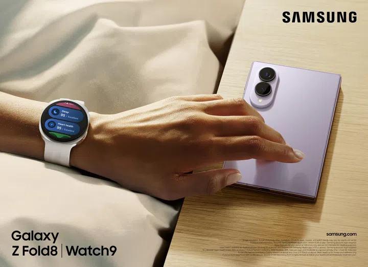 Samsung Galaxy Z Fold8与Galaxy Watch9情境展示图。一只佩戴白色Galaxy Watch9的手轻放在Lavender（薰衣草紫）配色的Galaxy Z Fold8上，手机以折叠状态放置于木质桌面。Galaxy Watch9圆形表盘显示Samsung Health健康数据，包括睡眠评分95分（Excellent）及心脏健康评分95分（Excellent），展现健康监测功能。Galaxy Z Fold8背面采用雾面淡紫色机身设计，配备双镜头相机模组，整体外观简约精致。背景为柔和米白色布料与暖色自然光，营造舒适生活氛围，呈现Galaxy Z Fold8与Galaxy Watch9搭配使用时，在日常生活、健康管理及Galaxy生态体验中的应用场景。
