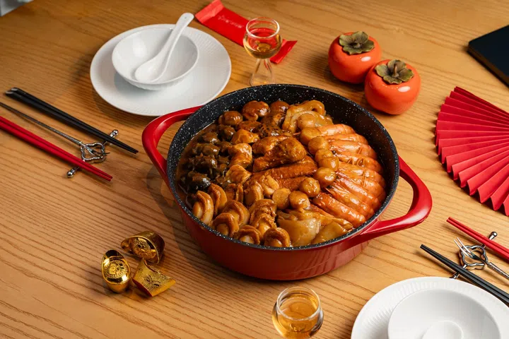 红色双耳锅中盛装福建风味盆菜，整齐铺排整虾、鲍鱼、海参、花菇与多款海味与肉类，汤汁浓亮入味，摆放在木质餐桌上，周围搭配红色新年装饰、金元宝摆件、酒杯与餐具，营造温馨喜庆的农历新年团圆饭氛围，新加坡盆菜推荐2026 农历新年必吃餐厅与价格整理。