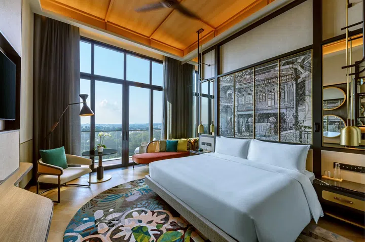 Artyzen Singapore Deluxe Balcony King客房，采用现代雅致设计，配备一张特大双人床、大面积落地玻璃窗及私人阳台，可俯瞰绿意景观。房内以木质元素和暖色灯光营造温馨氛围，设有休闲沙发、单人座椅、小圆桌及现代吊灯，床头背景饰有新加坡传统店屋风格艺术壁画，融合现代奢华与本地文化特色，营造舒适惬意的城市度假体验。