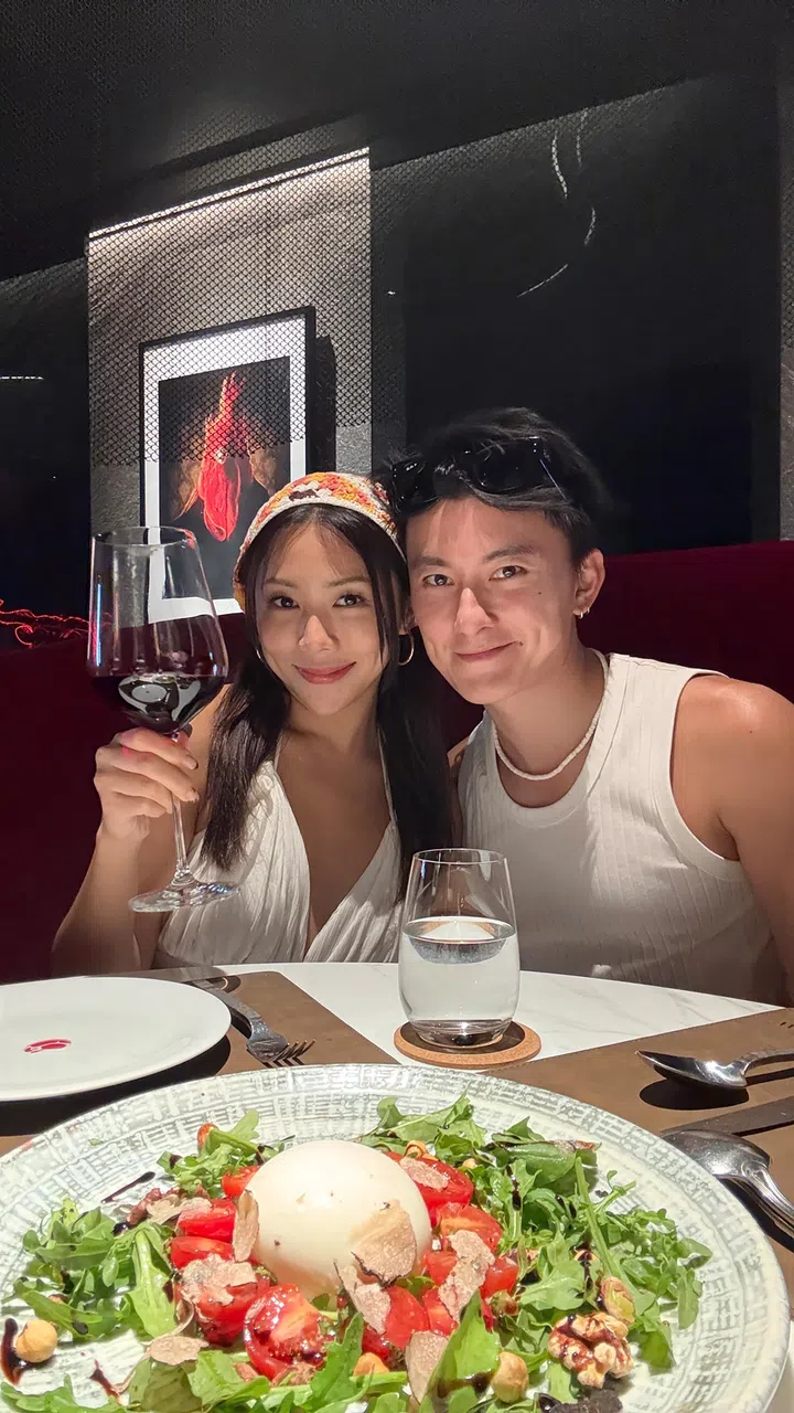 Edwin Goh与Rachel Wan在餐厅里并肩合影，两人对着镜头微笑，Rachel手举红酒杯庆祝，桌上摆放精致沙拉与餐具，营造浪漫温馨的约会氛围。Rachel Wan：最好的感情，是让我更像自己