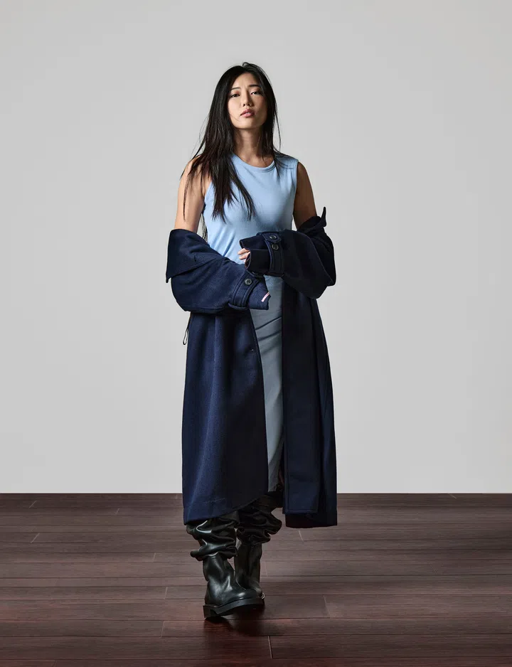一名长发女性站在浅灰背景前，身穿浅蓝色无袖上衣、深蓝长外套与深色长靴，双手轻握外套，姿态自然自信，脚下是深木色地板。