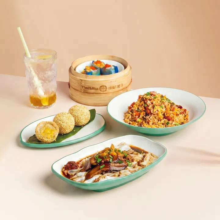 浅色背景前摆放多道 Tim Ho Wan 国庆限定港点，包括肉脞肠粉、培根午餐肉 XO 炒饭、芋泥金沙煎堆，以及蓝色外皮的叻沙海鲜饺，旁边搭配一杯清爽饮品。整体以简洁明亮的风格呈现，将新加坡本地风味与港式点心结合。