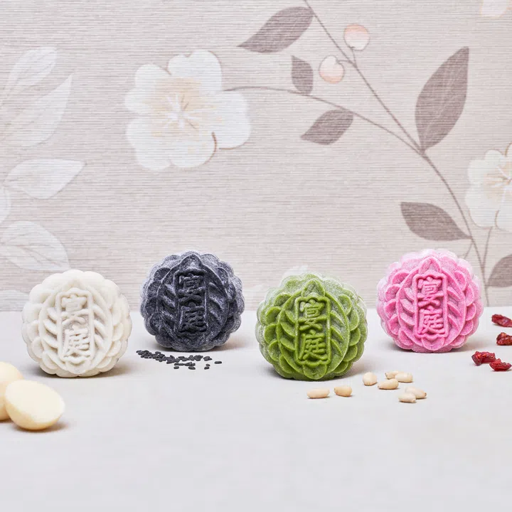 四枚不同颜色的迷你冰皮月饼（白色、黑色、绿色与粉红色）整齐摆放在浅色桌面上，饼面压印“宴庭”字样。背景为米色花卉壁纸，桌面散落着白巧克力、黑芝麻、松子与蔓越莓干，点缀出食材元素。The St. Regis Singapore， 2025中秋月饼必买推荐