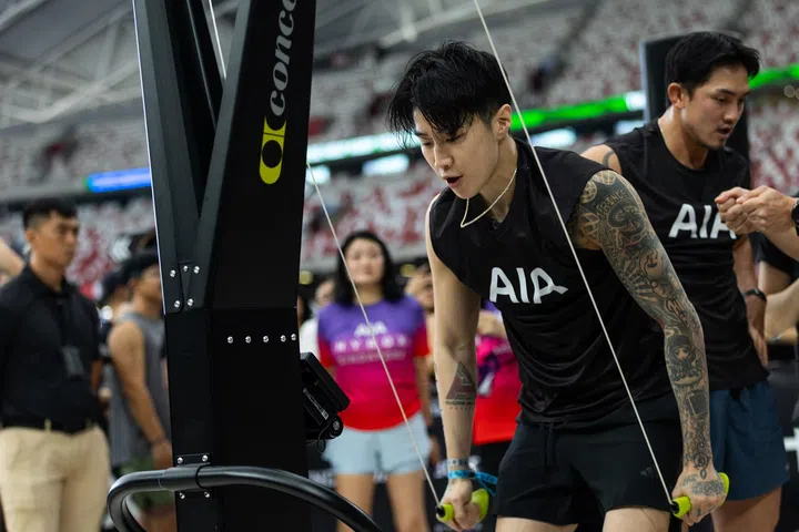 Jay Park在HYROX比赛中使用SkiErg训练设备进行高强度拉动动作，神情专注、肌肉紧绷，身穿AIA运动背心，现场观众在背景围观加油，比赛气氛紧张热烈。