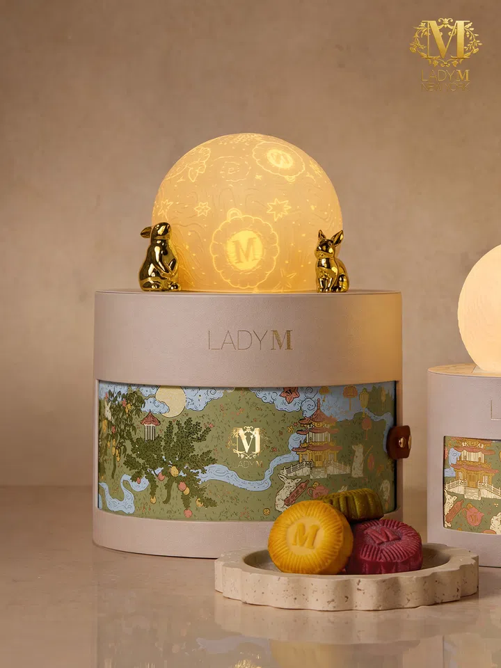Lady M 2026中秋“Moonlit Reverie”月饼礼盒场景图，画面中央为浅米色圆筒造型礼盒，盒身饰有中式园林、亭台、树木与月夜插画，并印有金色Lady M标志。礼盒顶部放置发光的月球造型灯饰，两侧点缀金色兔子摆件；前景陈列黄色、绿色与紫红色月饼，其中一颗印有Lady M字母“M”。整体以暖金色灯光呈现优雅梦幻的中秋送礼氛围。