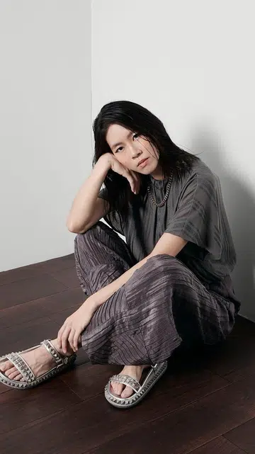 一张《女友》The Millennial Talk人物专题视觉图。画面左侧是一位留着及肩黑色长发的女生坐在白色墙角与深色木地板交接处，身体微微蜷曲，一只手托着脸侧，姿态放松自然，神情平静而若有所思地看向镜头。她身穿灰色宽松上衣，搭配深灰紫色压褶宽裤，并佩戴银色链条项链，脚穿银白色铆钉细节凉鞋。整体穿搭以低饱和灰调为主，呈现简约、中性且带有随性感的风格。人物背后形成柔和阴影，为画面增添层次与情绪感。
画面右侧为粉色背景，并以黑色大字写上“The Millennial Talk × Tan Siyou”，下方附有《女友》NUYOU标志。整体视觉风格简洁利落，同时带有安静、内敛且富有故事感的人物专题氛围。