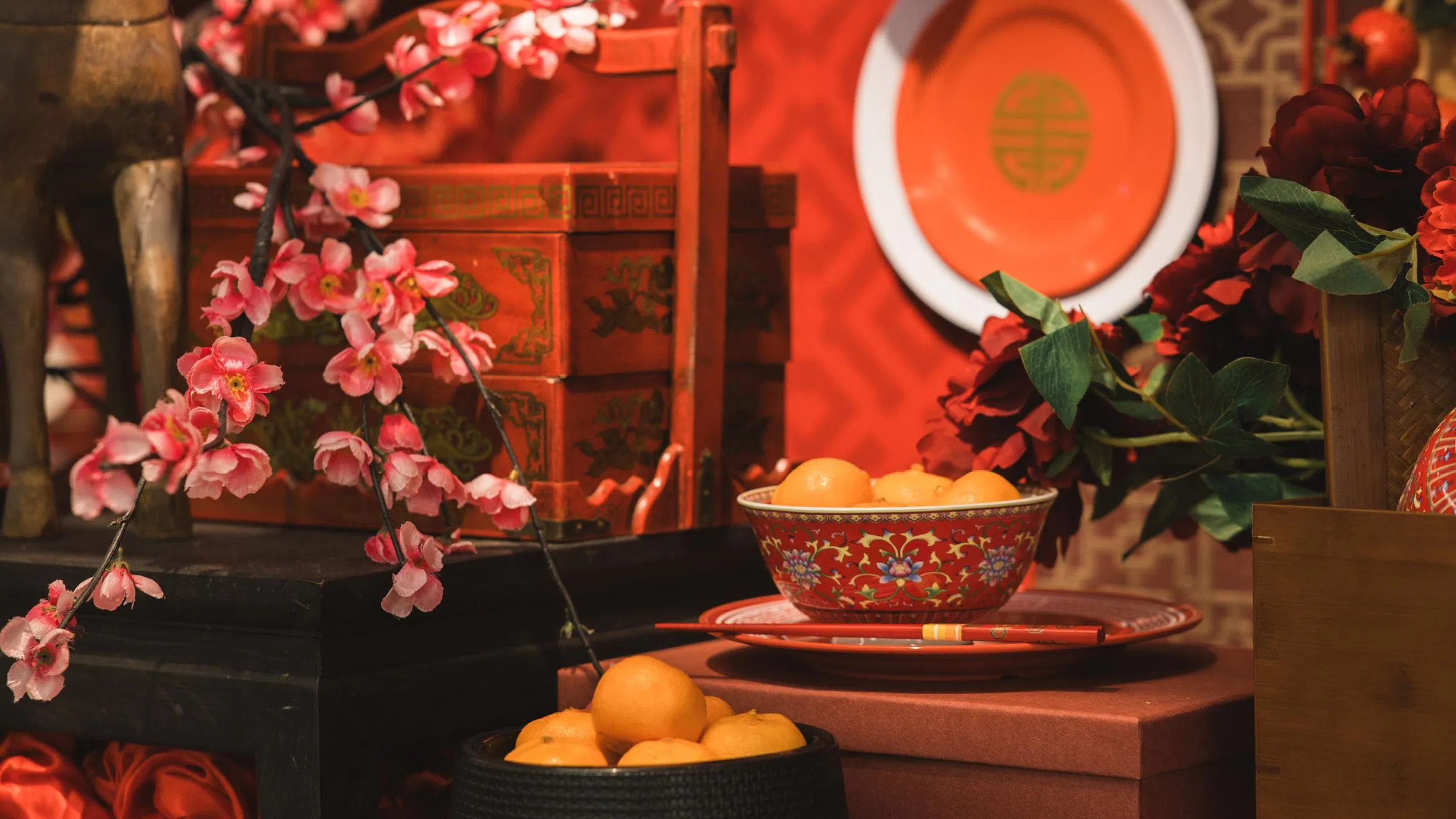 农历新年室内摆设场景，以红色为主调的节庆布置中，桌面陈列着盛满柑橘的红色花纹瓷碗与红色筷子，象征吉祥与丰收；背景可见传统红色木箱、圆形红盘与中式纹样装饰，搭配粉色梅花枝与红色花卉点缀，整体呈现出融合传统元素与精致美感的新春摆设氛围。