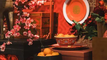 农历新年室内摆设场景，以红色为主调的节庆布置中，桌面陈列着盛满柑橘的红色花纹瓷碗与红色筷子，象征吉祥与丰收；背景可见传统红色木箱、圆形红盘与中式纹样装饰，搭配粉色梅花枝与红色花卉点缀，整体呈现出融合传统元素与精致美感的新春摆设氛围。