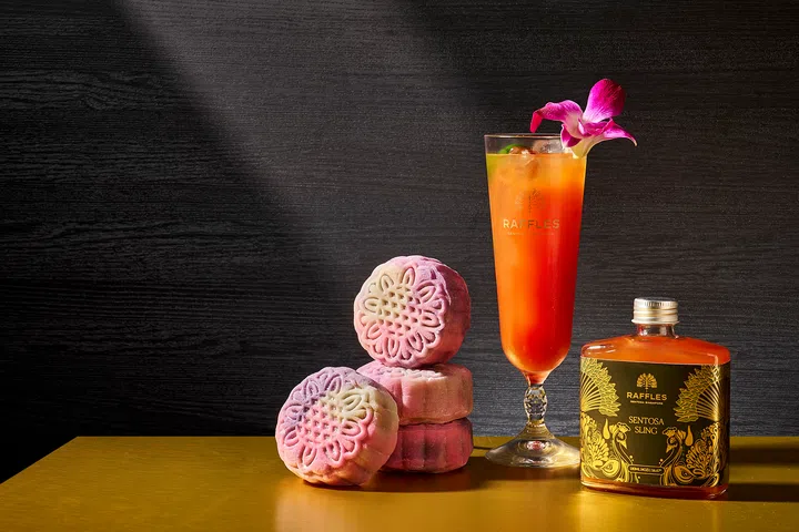三枚渐层粉紫色的冰皮月饼堆叠摆放，旁边是一杯高脚杯盛装的橙红色鸡尾酒，上方点缀紫色兰花。桌面还放置一瓶印有金色花卉图案的 Raffles Sentosa Sling 调酒，背景为深色木质纹理，整体氛围优雅而现代。Raffles Sentosa Singapore，2025中秋月饼必买推荐