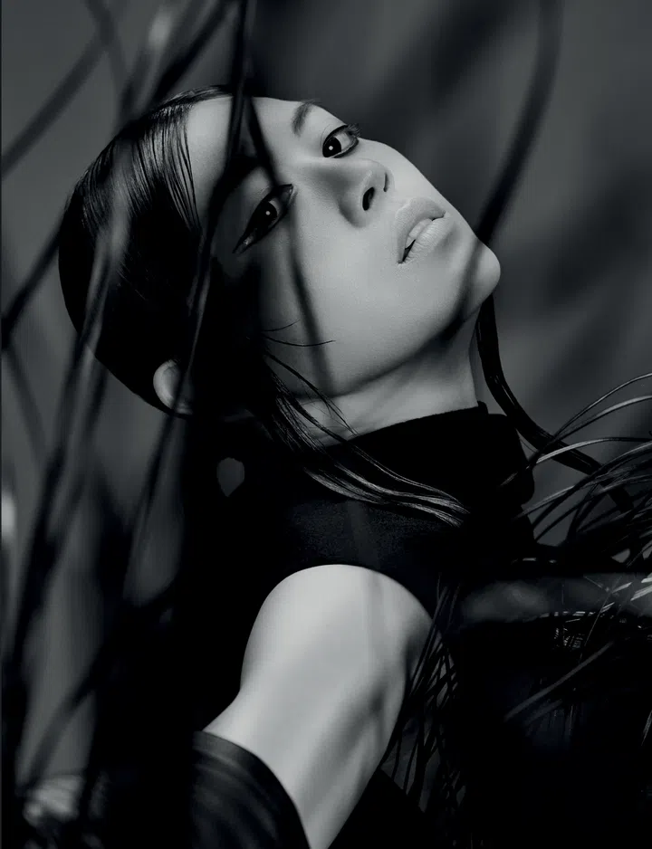 Jasmine Sokko穿着Sportmax连身裙，搭配长手套。她仰头凝视镜头，面庞被连身裙的流苏交织环绕，氛围神秘而时尚。