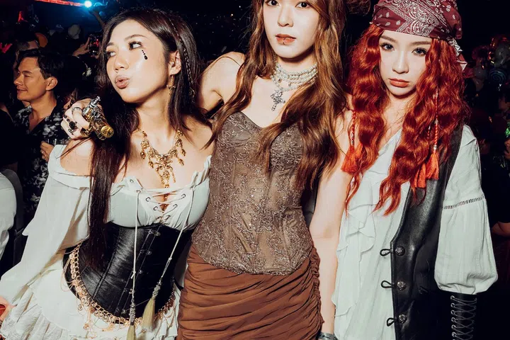 新加坡 Zouk 夜店万圣节派对现场照片，三名年轻女子穿着不同风格的万圣节服装合影。左侧女子身穿白色露肩上衣、黑色束腰及渔网丝袜，搭配金色项链与脸部装饰；中间女子戴着深棕色帽子，穿着复古风紧身上衣及棕色短裙；右侧女子则以红色长卷发、印花头巾、白色衬衫与黑色马甲打造海盗造型。背景可见昏暗灯光与参加派对的人群，展现 Zouk Halloween 的热闹气氛。