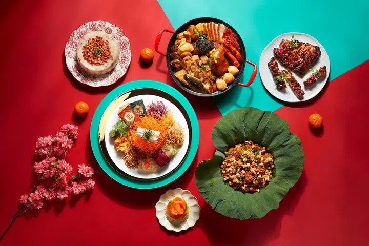 农历新年外带年菜组合展示图，红色与青绿色拼色背景上摆放多道中式佳肴，包括一盘色彩缤纷的捞起鱼生，搭配红萝卜丝、白萝卜丝、紫甘蓝、脆饼与酱包；一锅盛满鲍鱼、虾、带子、海参与卤肉的盆菜；以荷叶盛装的特色炒饭或炒料料理；一盘蜜汁烧排骨，以及甜点与年糕，整体呈现适合家庭团圆的新加坡农历新年外卖年菜与团圆套餐氛围。