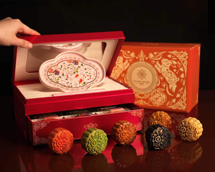 红金相间的 Old Seng Choong 月饼礼盒，打开后可见内附一只花卉纹饰的精美瓷盘，下层抽屉盛放月饼。前方摆放六枚不同颜色与口味的月饼，包括绿色、黑色、金黄色与焦糖色。背景为同系列的装饰礼盒，整体氛围喜庆大方。Old Seng Choong 老成昌，2025中秋月饼必买推荐