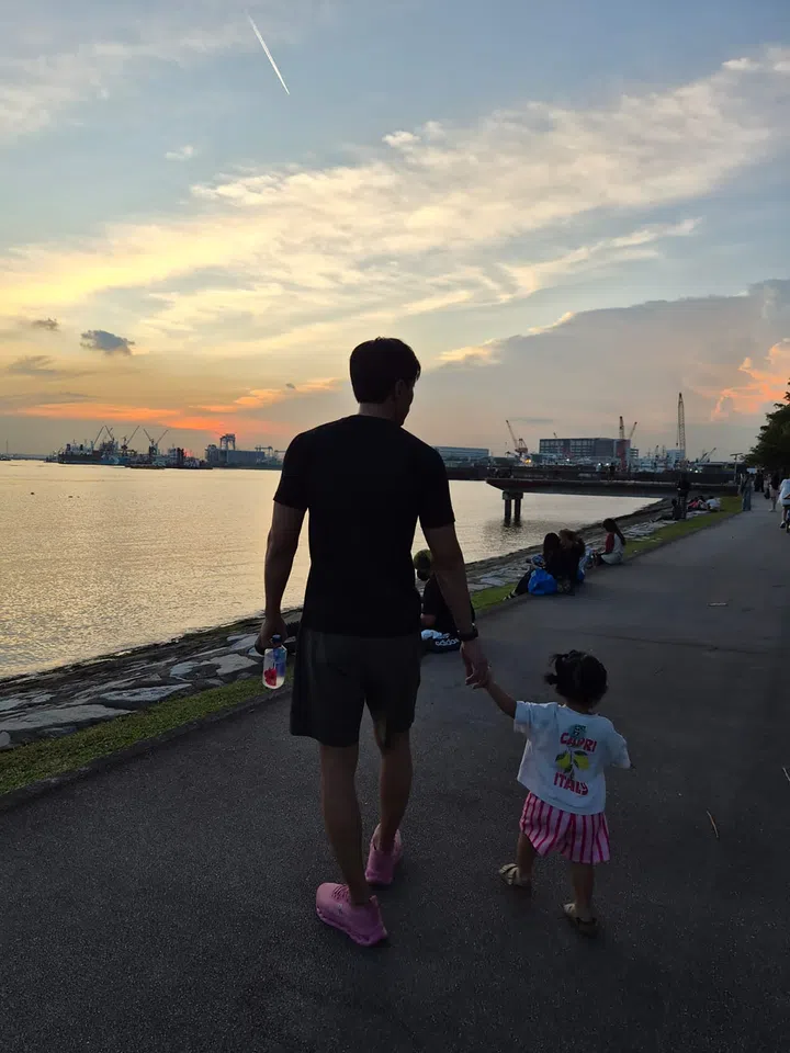 陈泂江身穿黑色上衣与短裤，脚踩粉色鞋，牵着穿粉白相间衣裙的女儿在海边步道散步。夕阳映照在水面上，背景有港口与远处行人，画面温馨宁静。陈泂江独家专访。