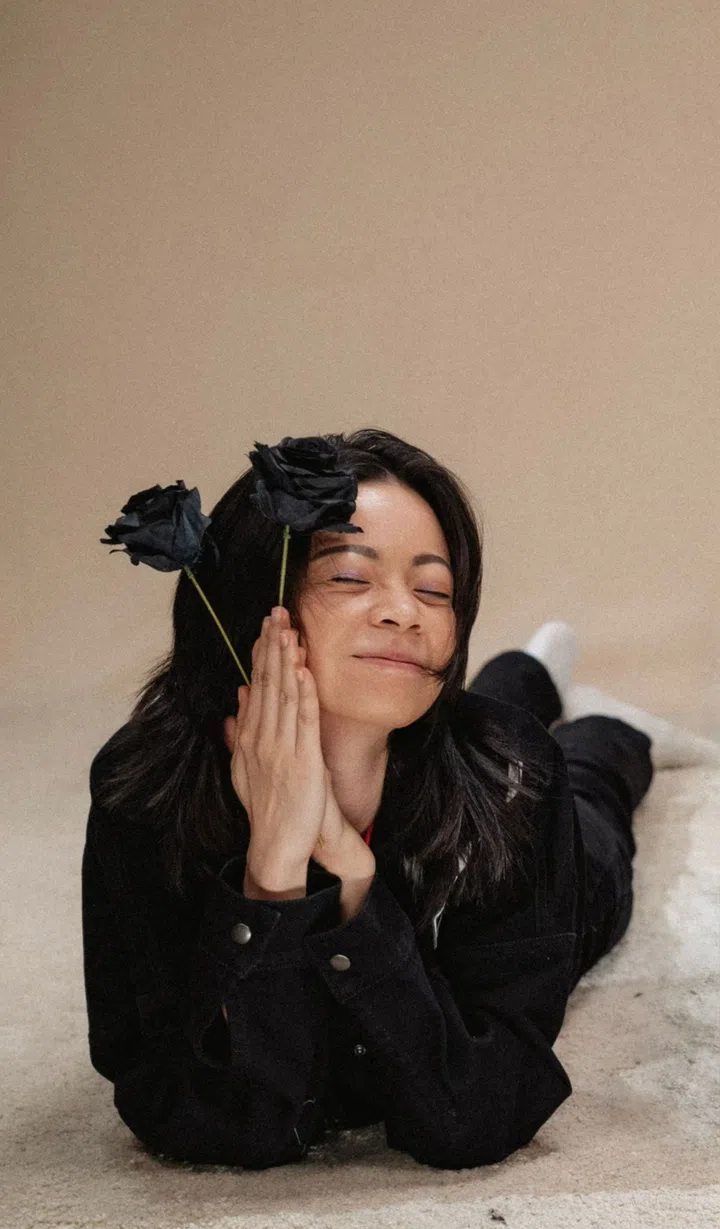 RAE SOH身穿黑色外套，趴在地毯上双手合十托脸，脸上带着轻松笑容，头侧别着两朵黑色玫瑰，呈现出俏皮又温柔的氛围。Half Open Tales新加坡流行庞克乐队女鼓手。