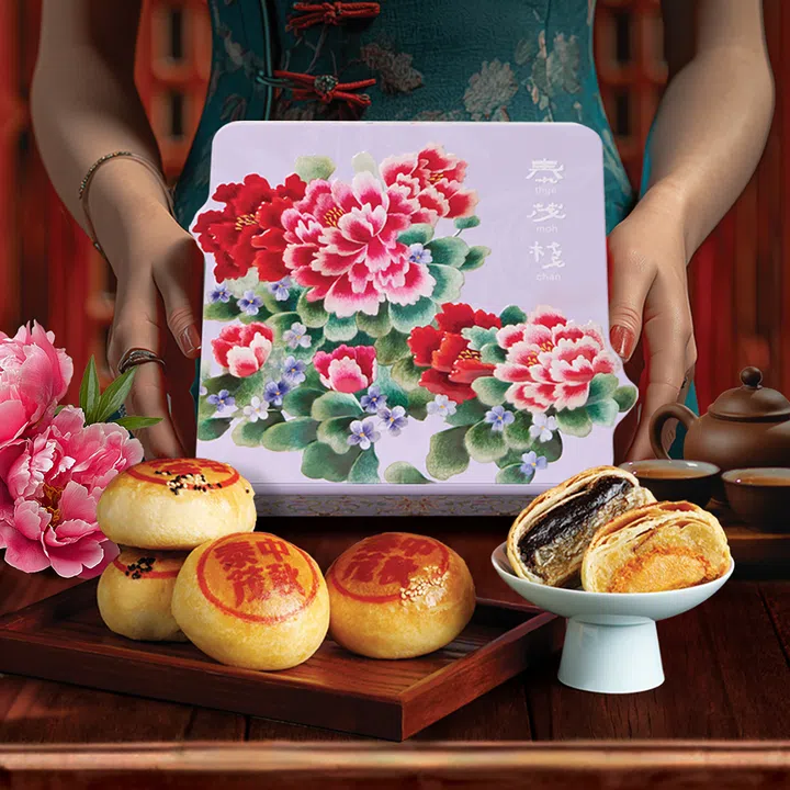 一名身穿旗袍的女子双手捧着一款以牡丹花为主图的紫色铁盒礼盒，盒上印有“Thye Moh Chan”字样。前方木盘上摆放数枚印有红色“中秋”字样的潮式饼，旁边白瓷碟内放置对半切开的饼，露出黑芝麻与蛋黄馅。桌面还点缀着茶具与牡丹花。Thye Moh Chan，2025中秋月饼必买推荐。