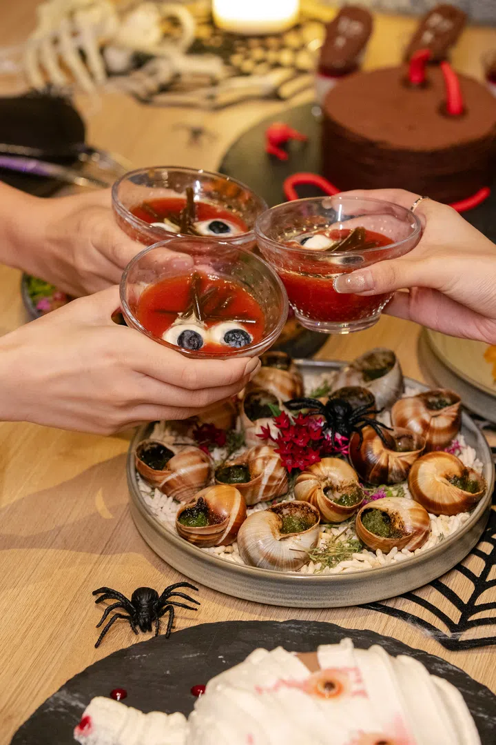 三人举起红色主题鸡尾酒庆祝，桌上摆放造型惊悚的蜗牛料理与万圣节甜点，营造节日氛围。新加坡万圣节去哪里玩推荐。