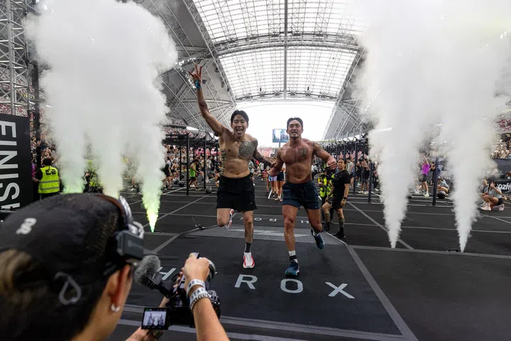 Jay Park在HYROX比赛冲过终点线，与搭档一同冲刺庆祝，双手举起、表情兴奋，现场喷烟效果与观众欢呼营造出热烈的完赛氛围。