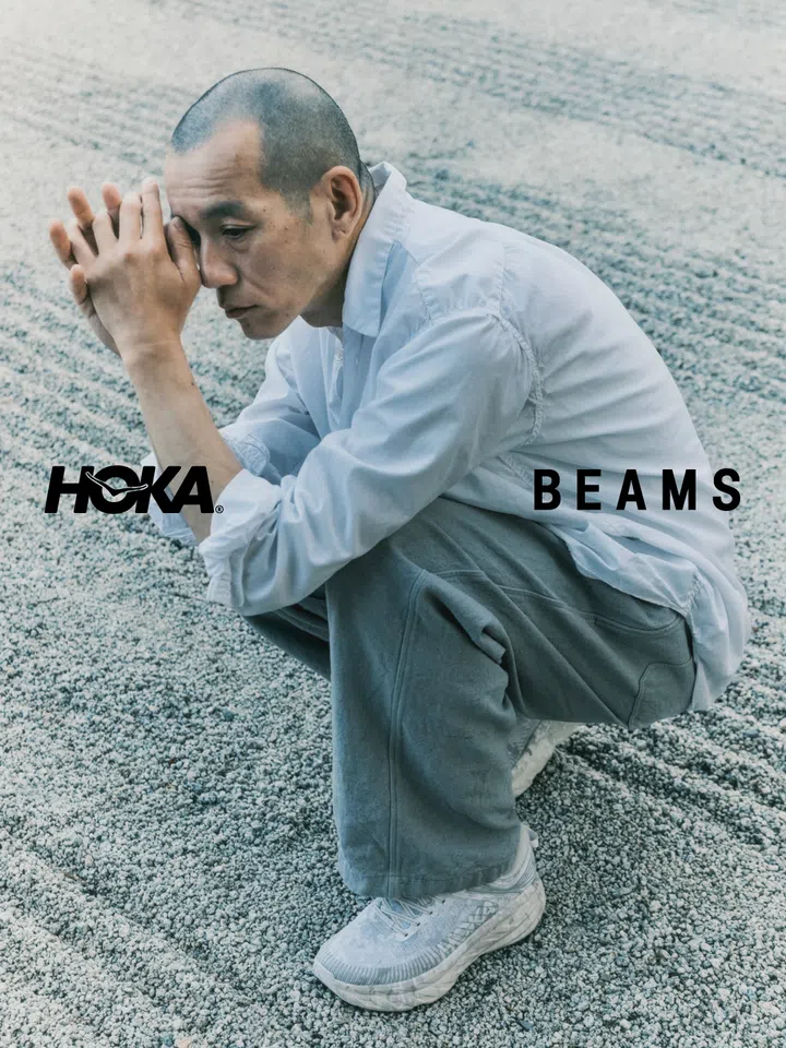 HOKA x BEAMS联名宣传照，一名剃着短发的男子身穿浅灰蓝色宽松衬衫和灰色长裤，蹲坐在铺满灰白色碎石的地面上，双手交握靠近脸侧，神情沉静。脚上穿着HOKA x BEAMS联名Bondi 7鞋款，鞋身以浅灰、米白等低饱和色调呈现，与碎石背景自然融合。画面整体采用偏冷的灰蓝色调，左下方印有黑色HOKA标志，右侧则是BEAMS字样，突出双方首次联名的简约街头风格。