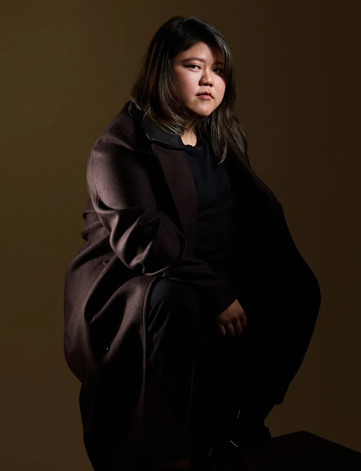 Jolinna Ang 坐在深色背景前，穿着深咖啡色外套与黑色内搭，在侧光下神情沉静而坚定，整体画面呈现柔和阴影与专注氛围。本地女性摄影指导Jolinna Angv