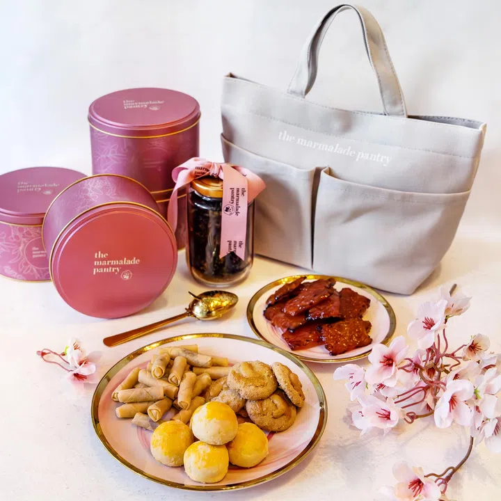 画面展示 The Marmalade Pantry 新春礼盒与年节点心组合陈列在浅色桌面上，整体风格温柔典雅，适合新春送礼场景。后方可见多款粉红色金属铁罐年饼礼盒，罐身印有 The Marmalade Pantry 品牌字样与细致花纹图案，右侧摆放一只浅灰色品牌手提托特包，适合搭配礼盒一同送出。中间摆放一瓶以粉色缎带装饰的瓶装茶饮或零食罐，旁边配有金色茶匙作为点缀。前景两只金边白盘分别盛放多款年节小吃，包括叁巴虾卷、腰果牛油曲奇、传统黄梨球与多片肉干，甜咸搭配丰富。画面右侧与前方点缀粉色樱花装饰枝条，营造柔和的新春氛围，整体呈现适合拜年与节庆送礼的年饼与礼盒组合选择。文章标题：“2026马年年饼推荐：新春送礼与拜年点心精选”。
